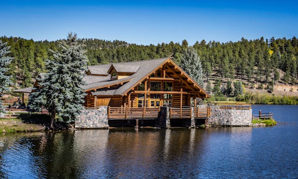 Beautiful wooden cabin by a serene lake surrounded by lush forest and clear blue sky.