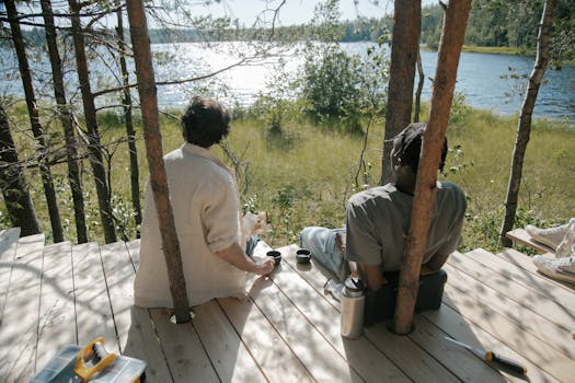 Two friends relaxing with coffee on a wooden dock by a lakeside, surrounded by nature, on a sunny day.